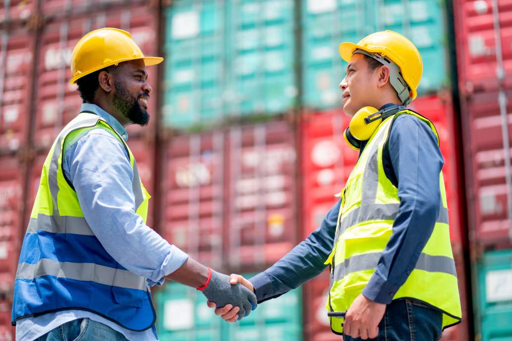 Afrikanischer amerikanischer Arbeiter oder Techniker sch&uuml;ttelt H&auml;nde mit asiatischen Mitarbeitern in der Arbeitsst&auml;tte Frachtcontainer