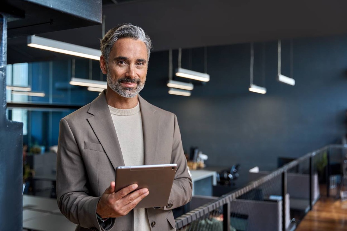 Confident mature businessman executive standing in office with tablet and doing digital banking