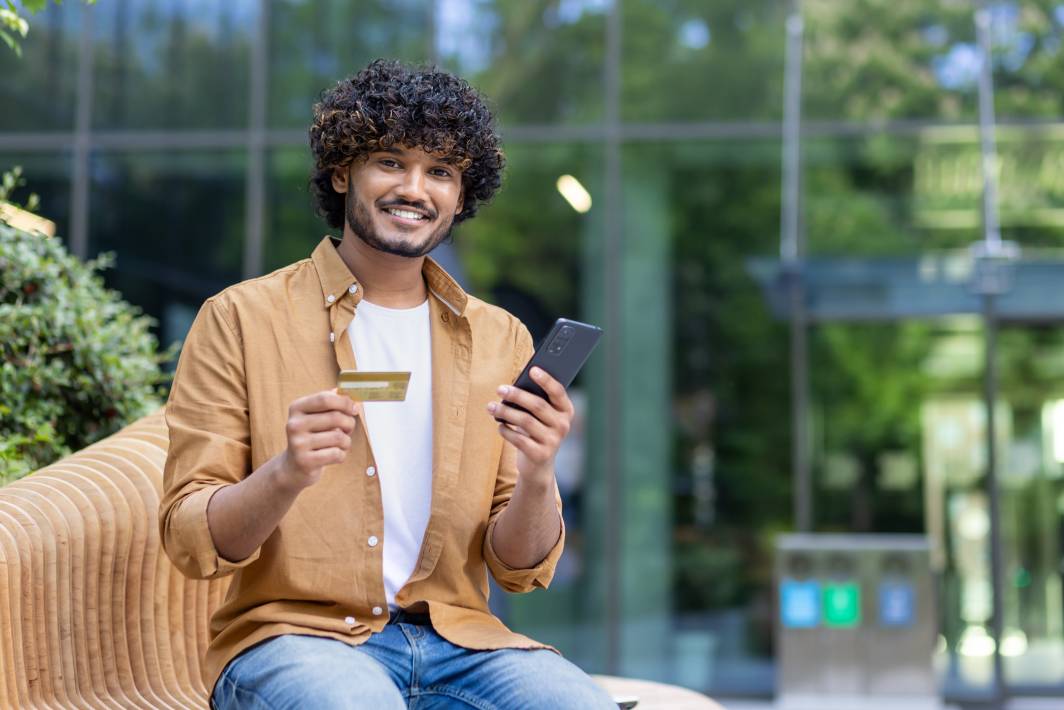 Portrait of a smiling young Indian man sitting outside on a bench, holding a credit card and a phone to transact through his offshore bank account